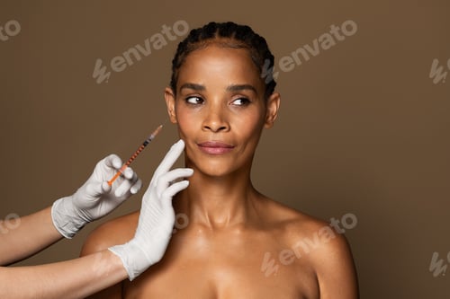Preview: Beautiful black middle aged woman getting facial injection in cheekbones zone, standing over brown