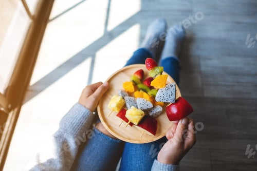 Preview: Top view image of a woman eating and holding a wooden plate of fresh mixed fruits on skewers