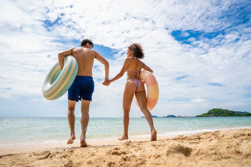 Preview: Rear view of a couple holding swimming inflatable and walk on the beach.