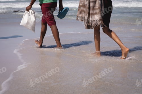 Preview: Low section of mixed-race couple walking on the waterside at beach in the sunshine
