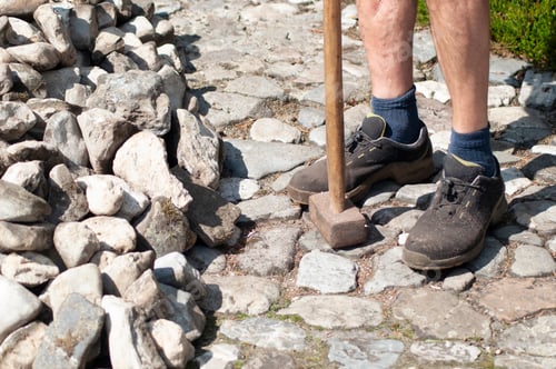 Preview: a gardener in dirty work clothes and boots holds a two-bladed iron hammer for working with stones