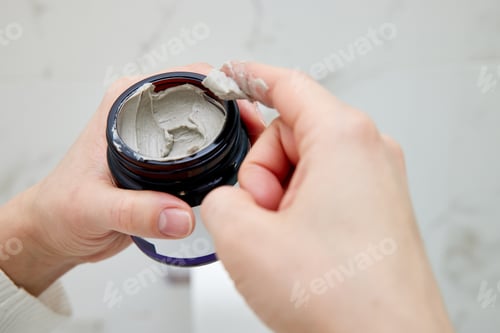 Preview: female hand takes a clay face mask from a jar.