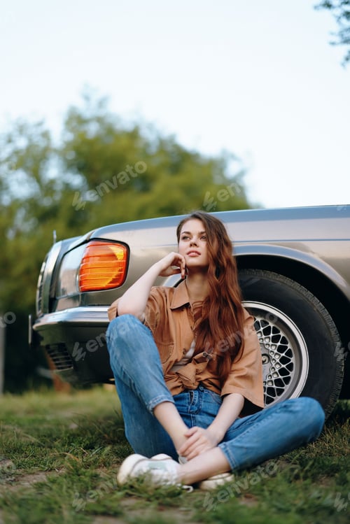 Preview: Young woman with long hair sitting on the grass next to a vintage car, wearing casual clothes