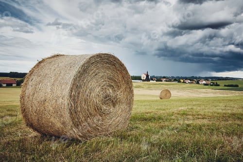 Preview: Straw bales stacked on field against landscape with village. Themes rural, agriculture, and weather.