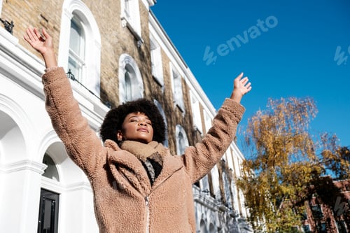 Preview: Young black woman enjoying the winter sun with arms up in the street.