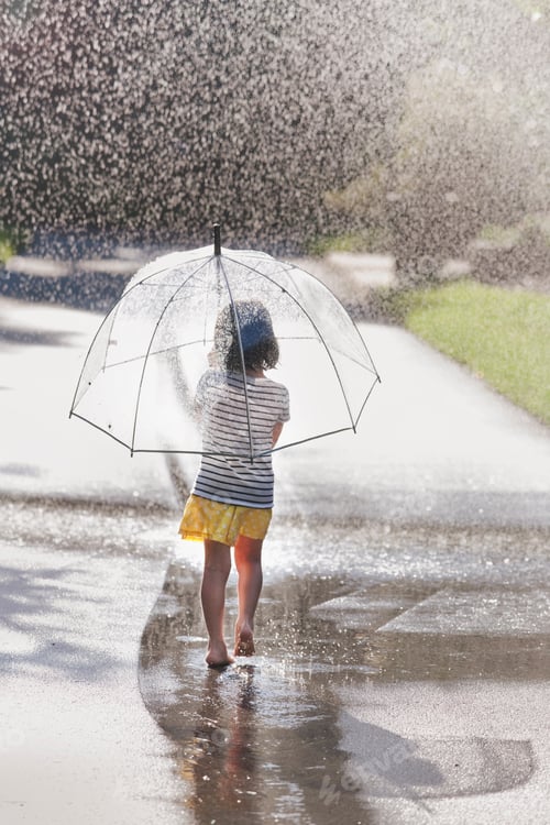 Preview: Rear view of barefoot girl carrying umbrella walking through street puddle