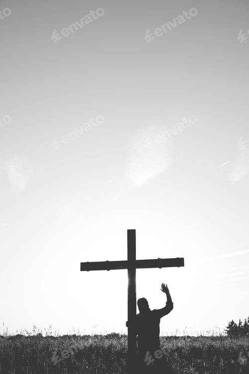 Vista previa: Vertical shot of a male standing near a cross with his hand up in black and white