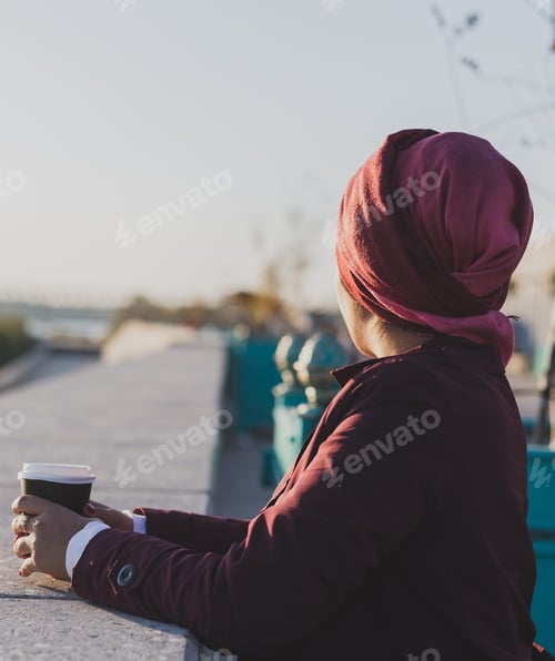 Preview: Young Arabic muslim woman in hijab standing at street seafront and drinking coffee take away - copy