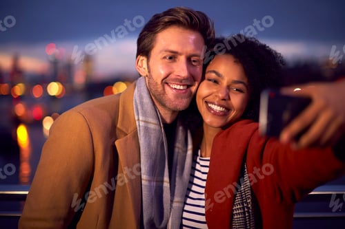 Preview: Couple Against City Lights Outdoors Wearing Coats And Scarves Posing For Selfie On Phone At Dusk