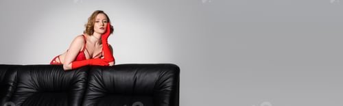 Preview: sensual young woman in red bra and gloves looking at camera near black couch isolated on grey,