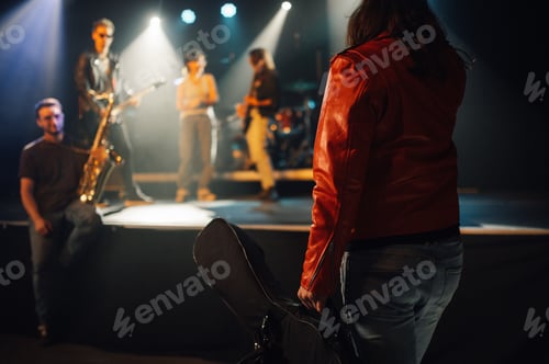 Preview: Roadie carrying guitar case walking towards stage with band performing