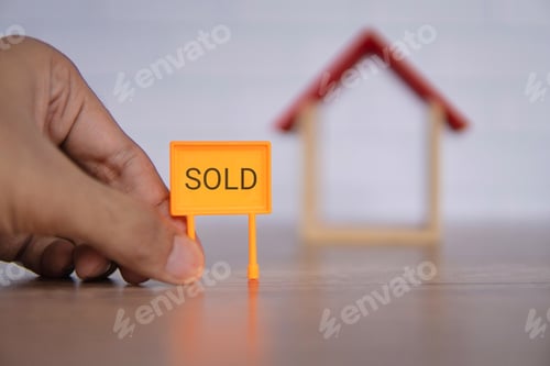 Preview: A close-up photo of a hand holding a bright orange "SOLD" sign in front of a miniature wooden house.