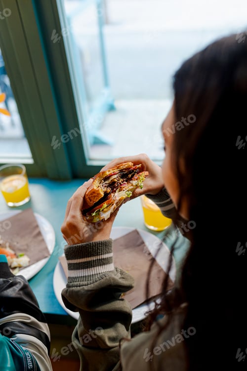 Preview: Young woman enjoying a delicious burger in a restaurant by the window