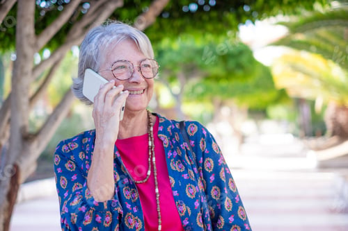 Preview: Cheerful senior woman in public park while using mobile phone enjoying tech and social