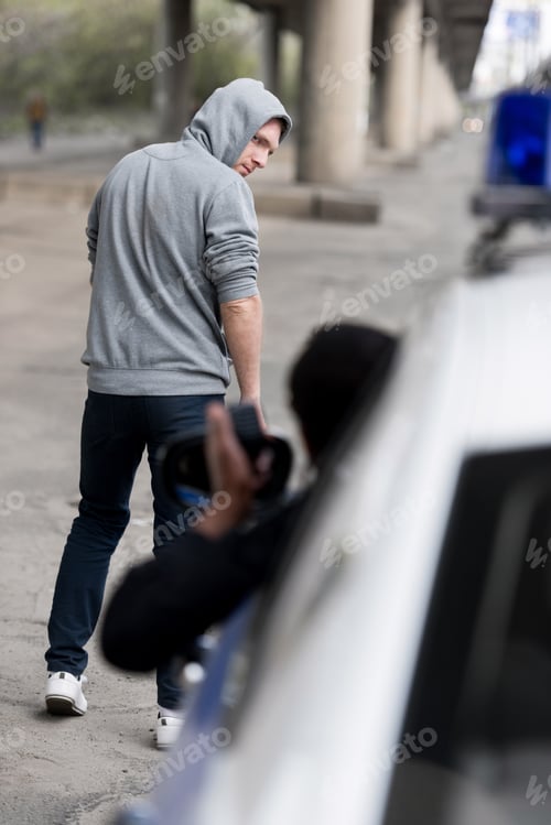 Preview: policeman looking on hooded young man walking away on street out of window