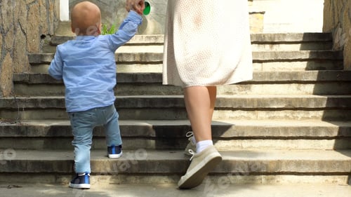 Preview: Close up of woman helping her little child boy to climb the stairs by holding his hand. Stock