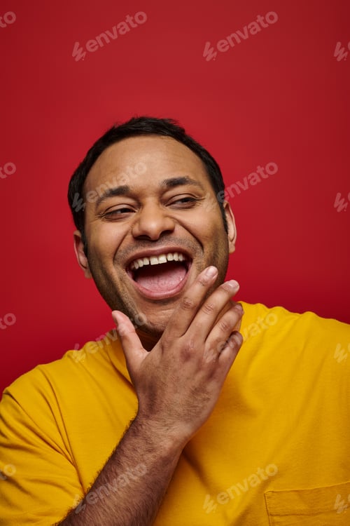 Preview: positive emotion, excited indian man in yellow t-shirt laughing with opened mouth on red background