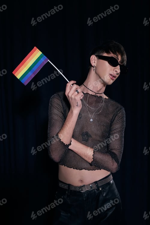 Preview: Young and fashionable queer person in sunglasses and shiny top holding lgbt flag while posing during