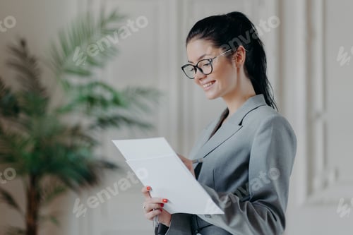 Preview: Glad female lawyer writes in corporate paper documents, wears optical glasses, formal clothes