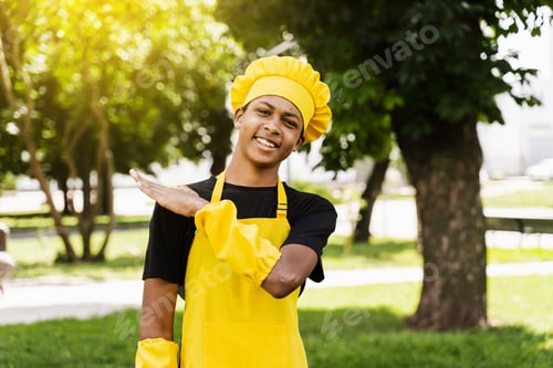 Preview: Happy African teen chef in yellow apron smiling outdoors, cafe promo image