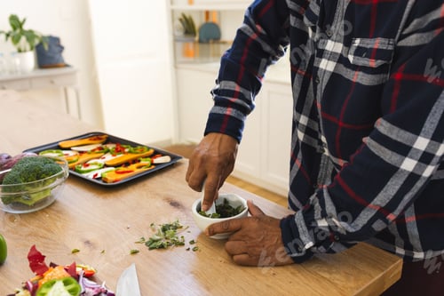Preview: Senior man wearing flannel shirt crushing herbs in bowl on kitchen countertop with tray, copy space