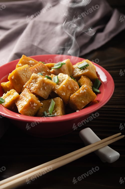 Preview: Crispy deep stir fried tofu cubes with chives in clay dish on wooden kitchen table with napkin aside