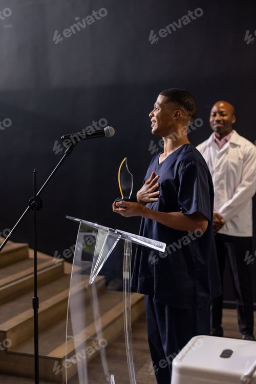 Preview: Man in scrubs holding award, speaking at podium, colleague smiling in background