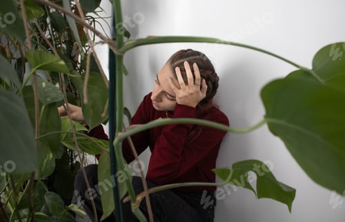 Preview: A girl is sitting in the corner by the wall in the room next to the houseplants. She's upset and sad
