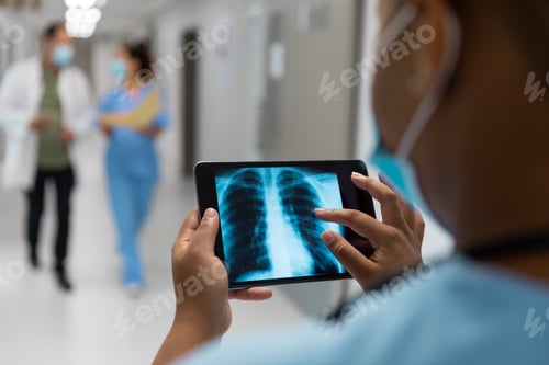 Preview: Mixed race doctor in corridor wearing face mask looking at lung x-ray on tablet