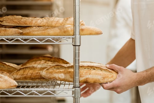 Preview: cropped shot of handsome baker putting fresh bread on shelf at baking manufacture