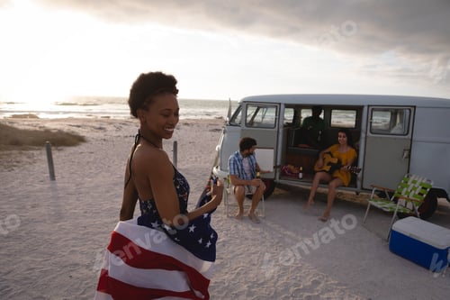 Preview: Woman holding an American flag against his friends sitting in a camper van at beach