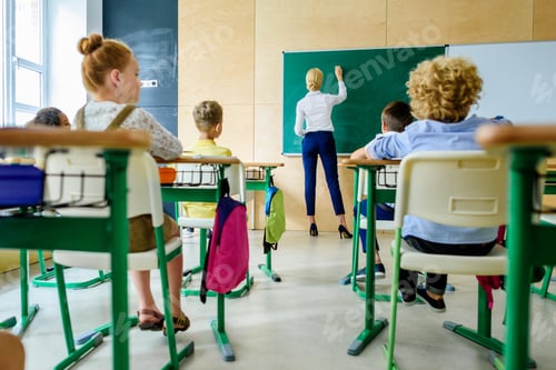 Preview: rear view of schoolchildren looking at teacher while she writing on chalkboard