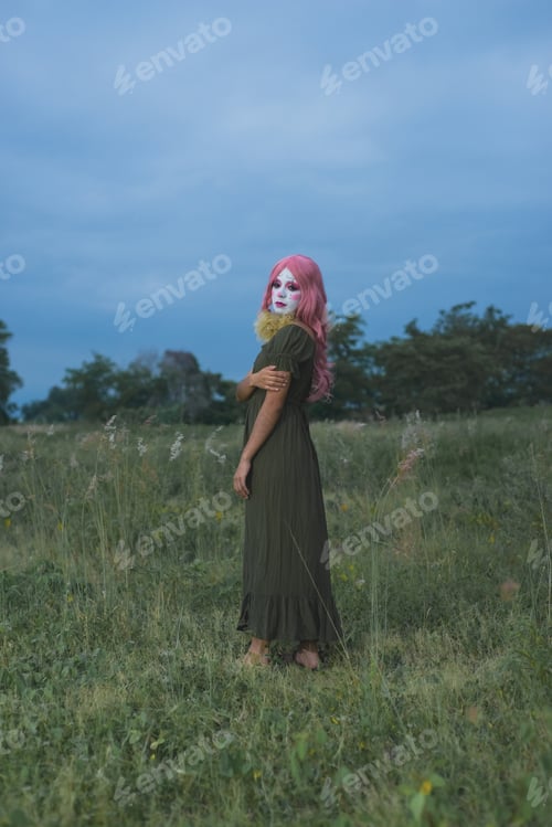 Preview: Young woman made up as a sad clown in a lonely field. Halloween makeup.