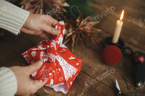 Preview: Hands wrapping christmas gift in red festive fabric on rustic table. Zero waste holiday, furoshiki