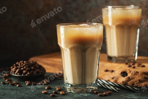 Preview: Ice coffee in a tall glass with cream poured over, ice cubes and beans on a dark concrete table.
