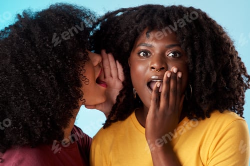 Preview: Studio shot of a young woman whispering in her friends ear against a blue background