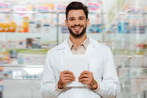 Preview: Druggist holding digital tablet and smiling at camera in apothecary