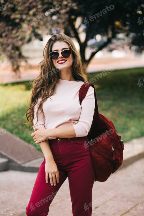 Preview: Cute girl with long hair in sunglasses with vinous bag and pants is smiling to camera in city park.