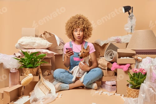 Preview: Stunned young woman with curly hair dressed casually poses with pet holds mobile phone finds apartme