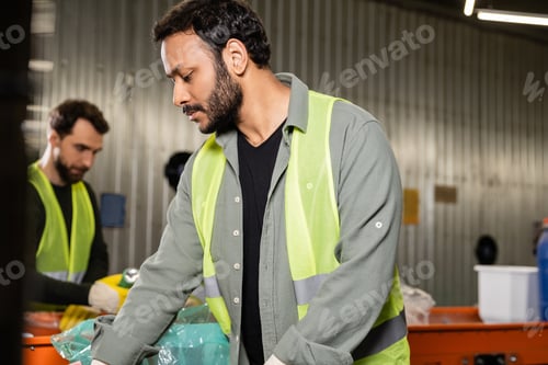Preview: Bearded indian worker in high visibility vest separating trash while working near blurred conveyor