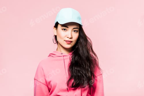 Preview: attractive asian woman in pink pullover and cap looking at camera isolated on pink