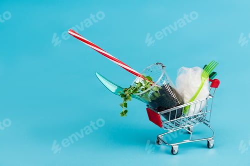 Preview: disposable plastic objects and plant in ground in small shopping cart on blue background