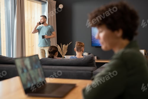 Preview: Young businessman standing by window, having drink and talking on mobile phone