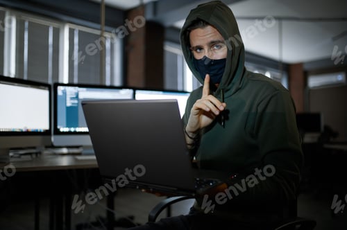 Preview: Male hacker in hood works on laptop in dark office