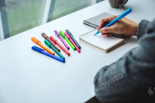 Preview: Closeup image of a woman writing on a blank notebook with colored pens on the table