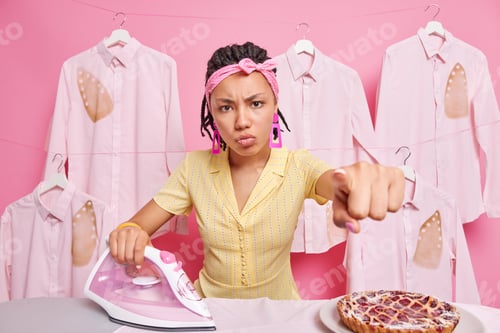Preview: Angry irritated housewife with dreadlocks wears headband points directly at camera has annoyed facia