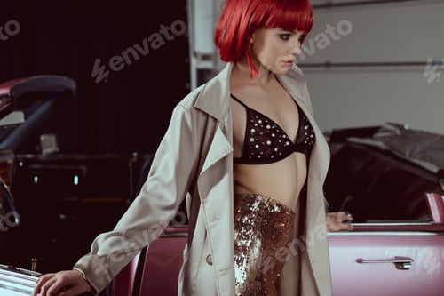 Preview: beautiful young woman in fashionable trench coat looking away while standing near retro car