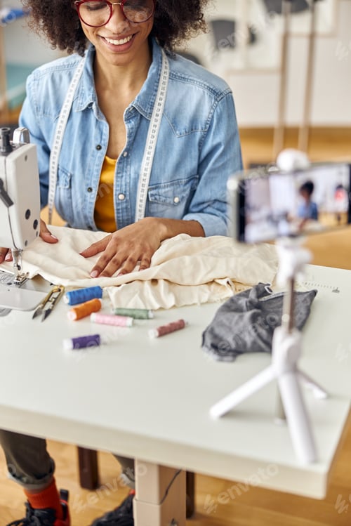 Preview: African-American lady designer sews with machine shooting video at table in studio