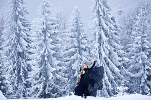Preview: Attractive woman in dark long fantasy outfit outdoors in deep snow on sunny winter day.