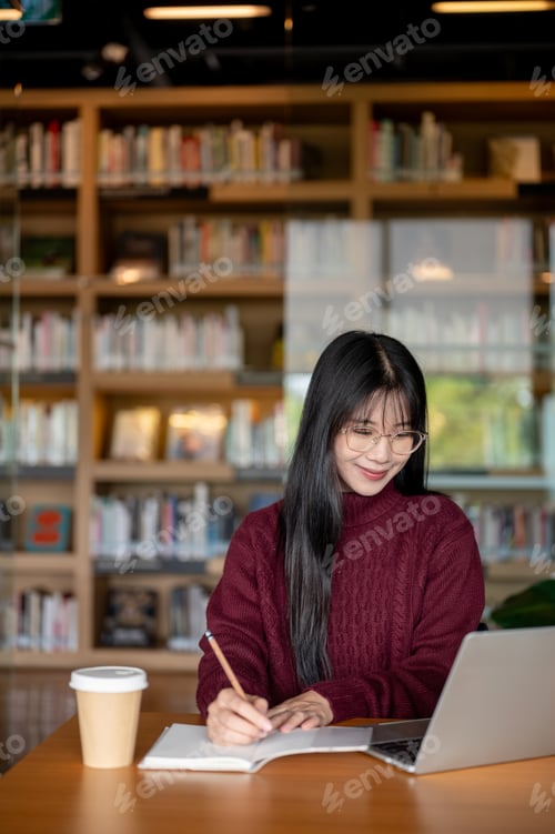 Preview: Smiling glasses asian woman writing in notebook as looking at laptop aside coffee at library's table